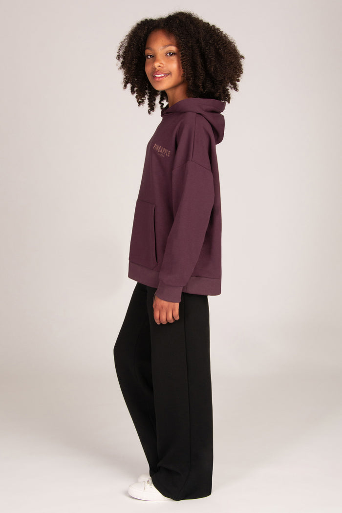 A young girl stands against a light grey studio background wearing a dark aubergine pullover hoodie, black wide-leg sweatpants, and white sneakers.