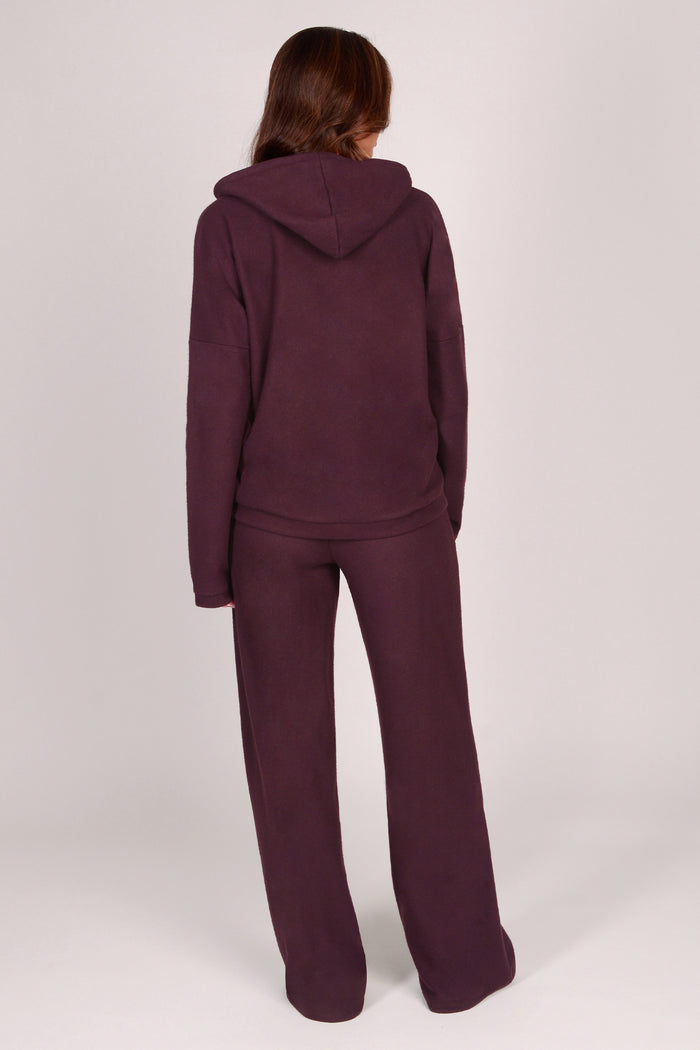 Woman wearing maroon brown wide leg trousers with matching dip hem hoodie on a plain background.