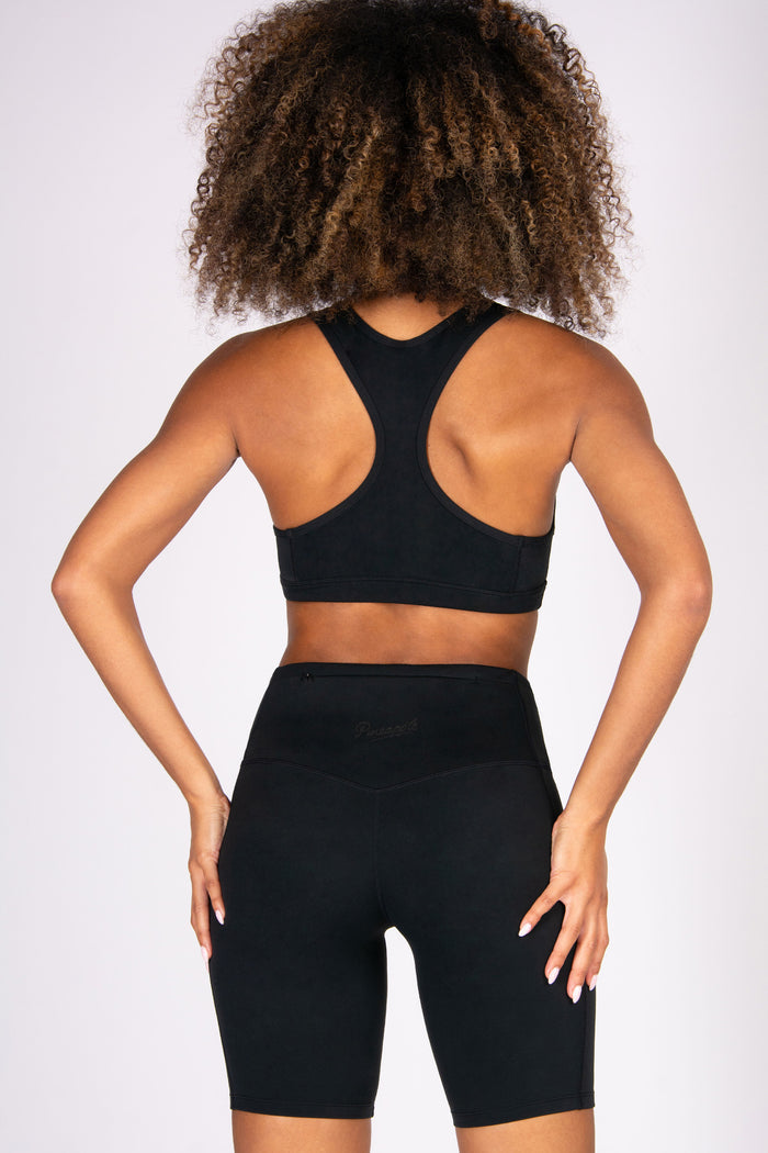 Woman wearing a black super soft racer back sports bra and matching cycling shorts with hidden zip pocket on a white background