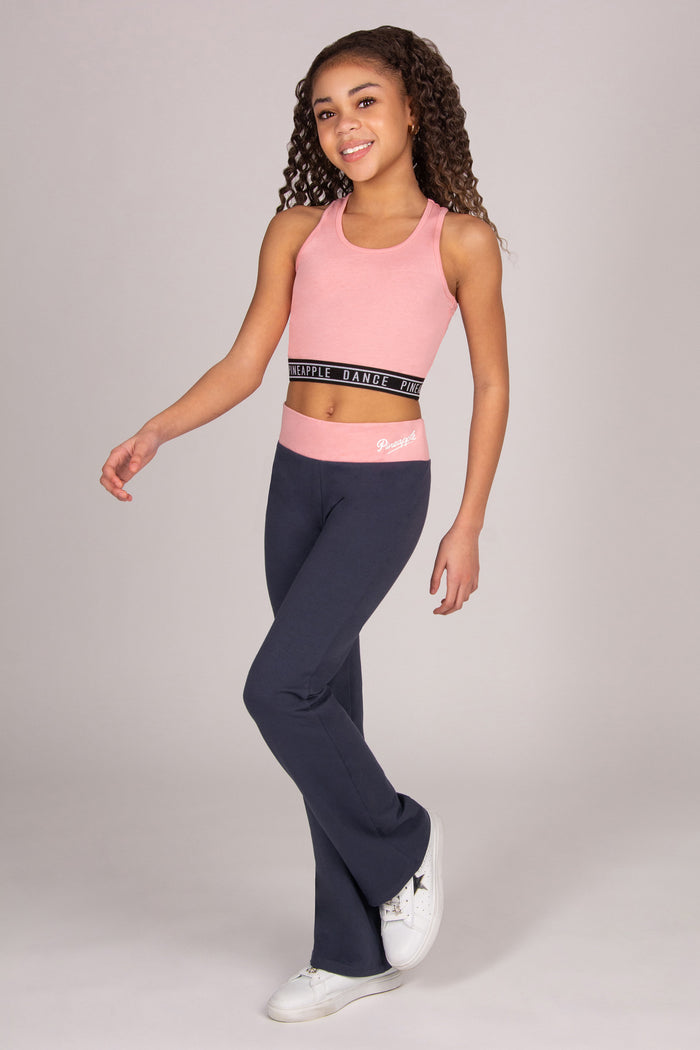 Girl wearing light pink sports crop top with matching dark grey bootcut trousers on plain background.