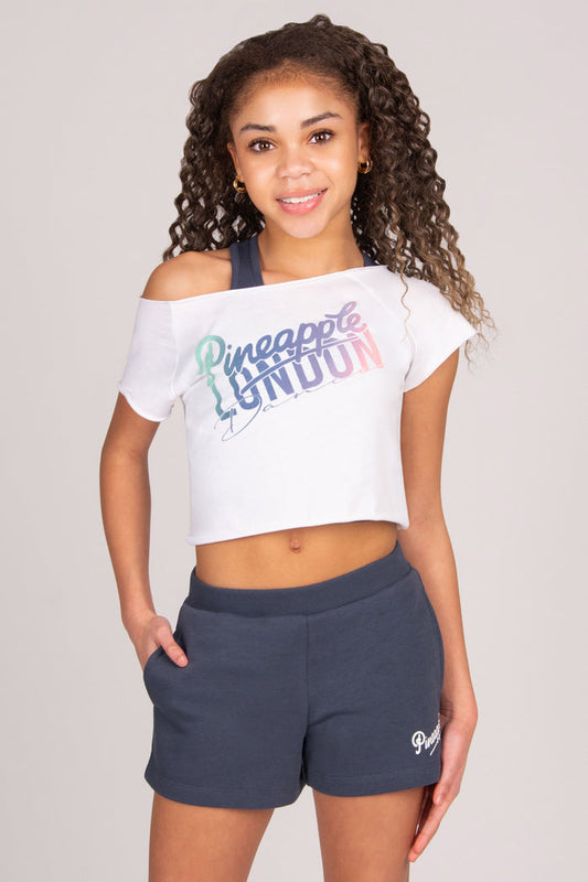 Girl wearing a white crop top with "Pineapple London Dance" print and dark grey shorts on a plain background