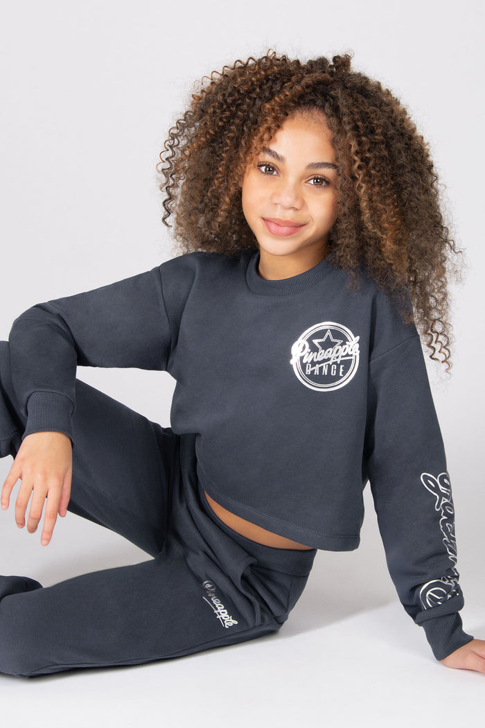 Young girl wearing a grey sweatshirt and matching trousers, sitting on a white backdrop.