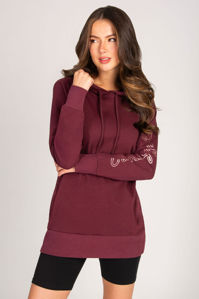 Woman wearing a burgundy hoodie with rose gold foil print on left sleeve on a plain background