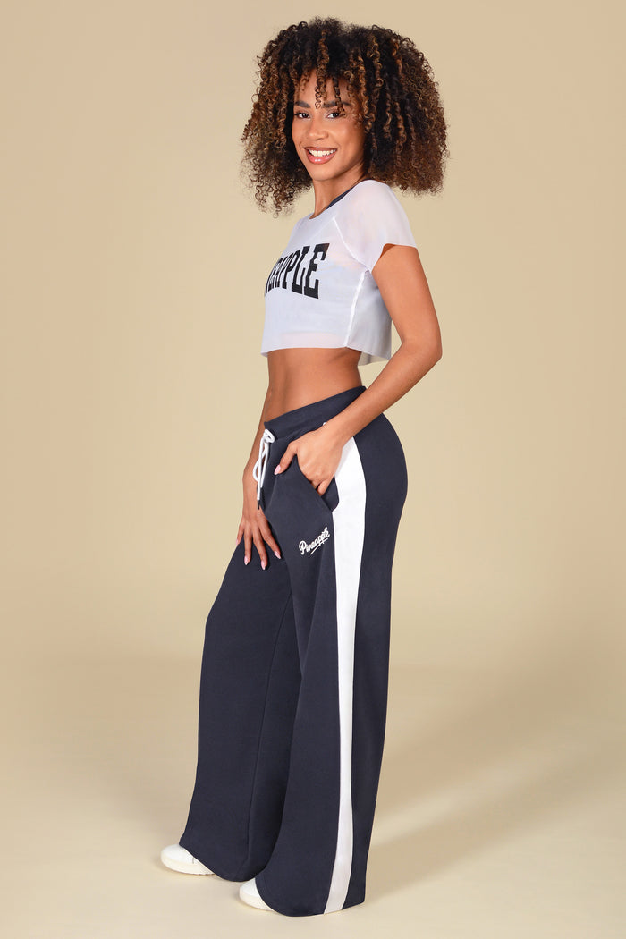 Woman wearing a white mesh crop top with 'Pineapple' logo paired with dark grey wide leg trousers against a light yellow background.
