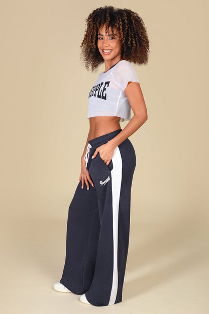 Woman wearing a white mesh crop top with 'Pineapple' logo paired with dark grey wide leg trousers against a light yellow background.