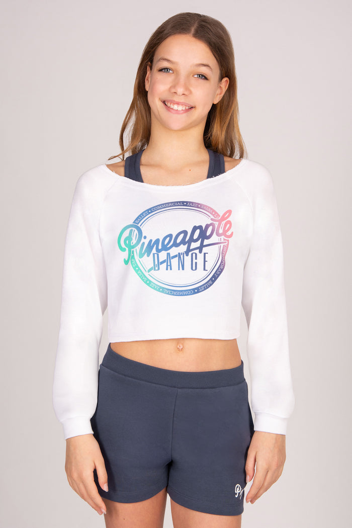 Girl wearing a white long sleeve crop top with ombre 'Pineapple Dance' logo and dark grey shorts on a plain background