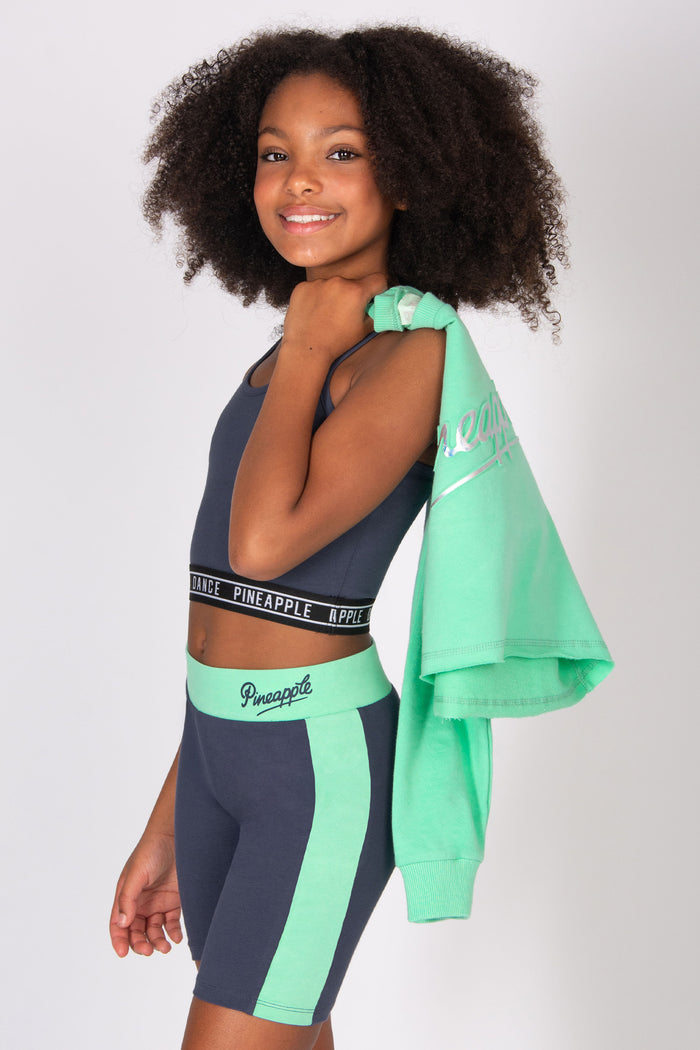Girl wearing a dark grey sports top and matching cycling shorts with a mint green sweatshirt over her shoulder on a white background