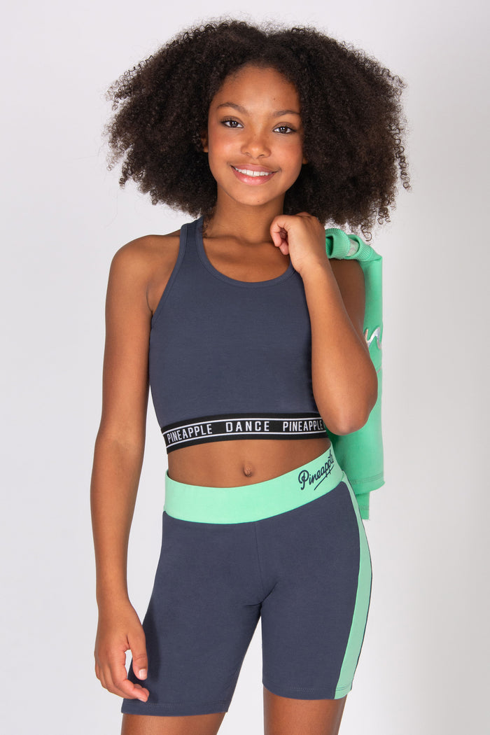 Girl wearing a dark grey sports top and matching cycling shorts with a mint green sweatshirt over her shoulder on a white background