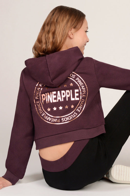 Girl sitting against a plain background wearing a maroon brown hoodie with rose gold 'Pineapple Dance Studios' design print on the back.