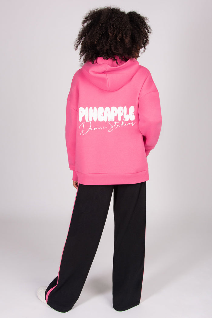 Girl wearing a pink oversized hoodie with 'Pineapple Dance Studios'  print on the back, standing against a white background.