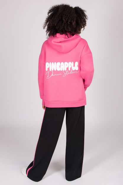 Girl wearing a pink oversized hoodie with 'Pineapple Dance Studios'  print on the back, standing against a white background.