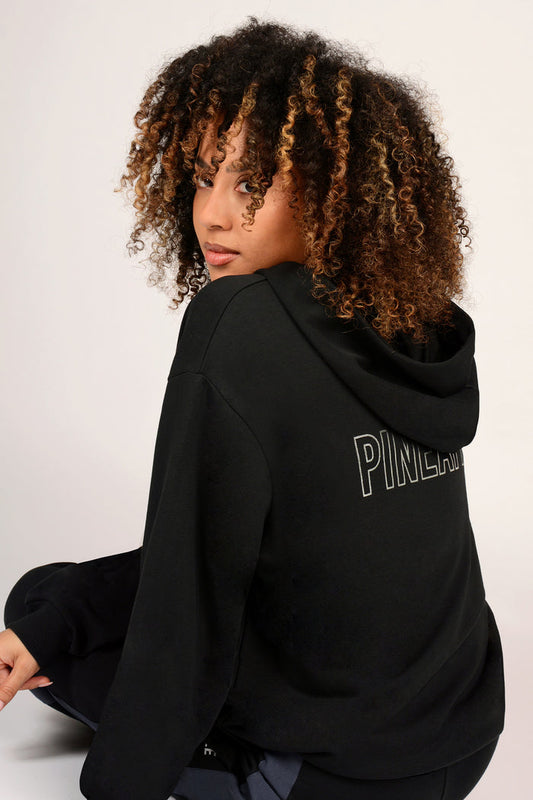 Woman wearing a black hoodie with 'PINEAPPLE' branding on the back, sitting against a white background.