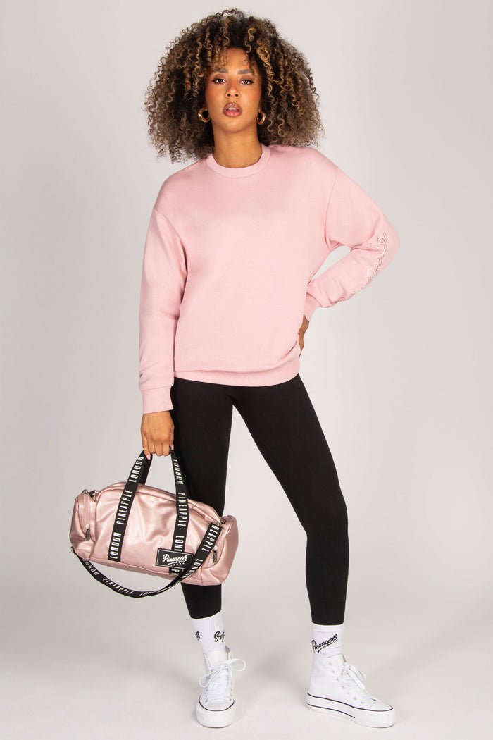 Woman wearing a pink sweatshirt, black leggings, and white sneakers holding a matching pink bag on a light gray background.
