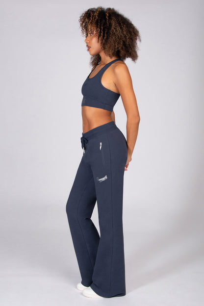 Woman wearing grey sports top and matching dark grey flared trousers with zip pockets on a light grey background.