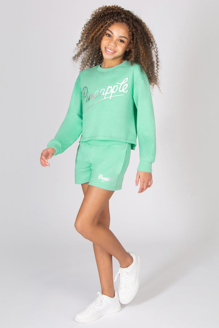 Girl wearing a mint green 'Pineapple' logo sweatshirt and sporty shorts set on a light grey background