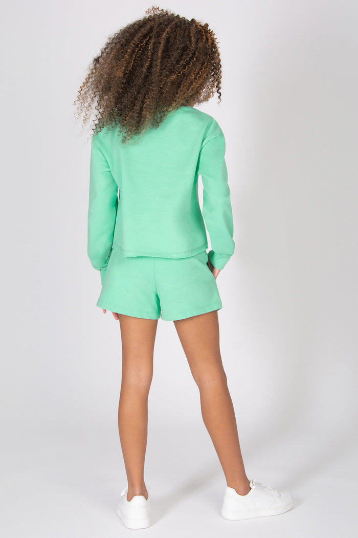 Girl wearing a mint green 'Pineapple' logo sweatshirt and sporty shorts set on a light grey background