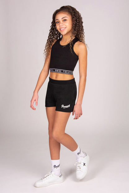 Young girl wearing a black athletic outfit with black pocket shorts on a plain background