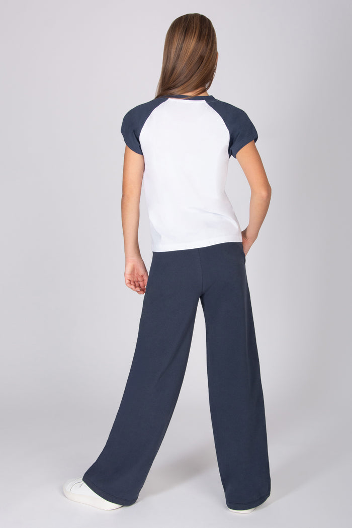 Teenage girl wearing a white t-shirt with contrast dark grey raglan sleeves with navy wide leg trousers on a light grey background