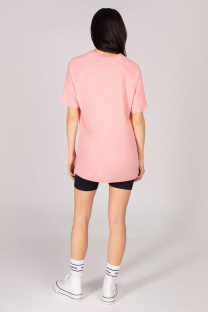 Woman wearing an oversized coral pink t-shirt with black cycling shorts and white shoes against a plain grey background.