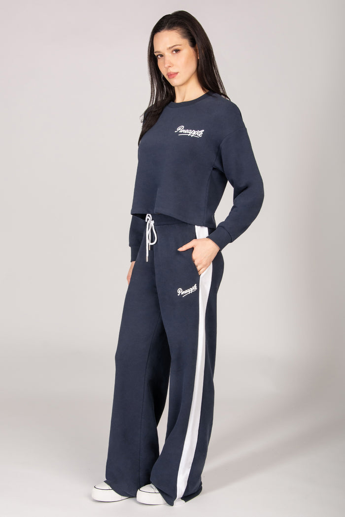 Woman wearing a dark grey midi sweatshirt with matching wide leg trousers with white side paneling on a plain background