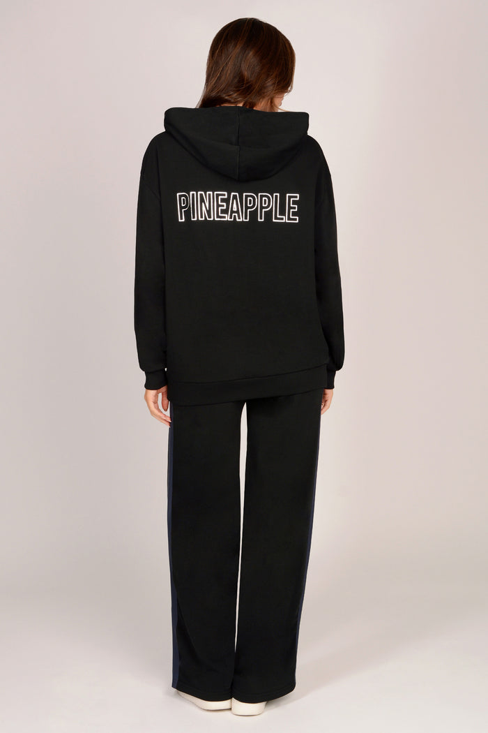Woman wearing a black hoodie with 'PINEAPPLE' text on the back, standing against a plain background.