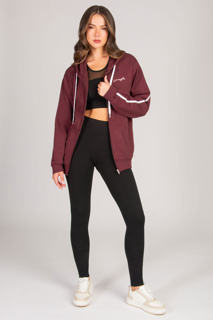 Woman wearing a maroon hoodie with zippers, pockets, and white drawcords and black leggings on a light grey background