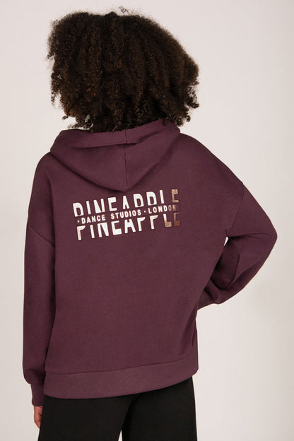 A girl shown from the back wearing a maroon oversized hoodie with 'PINEAPPLE DANCE STUDIOS - LONDON' printed in rose-gold across the back, paired with black trousers against a plain light background.