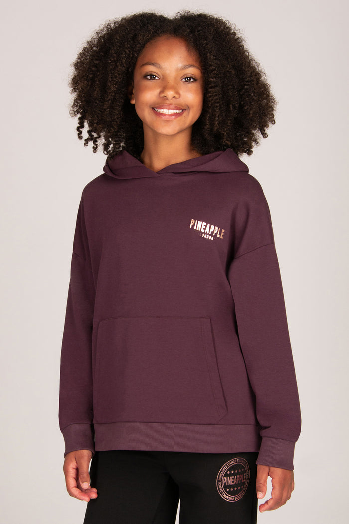 A smiling young girl wearing a dark plum purple pullover hoodie with a small rose-gold 'PINEAPPLE' logo on the left chest, paired with black trousers, against a solid light grey background.