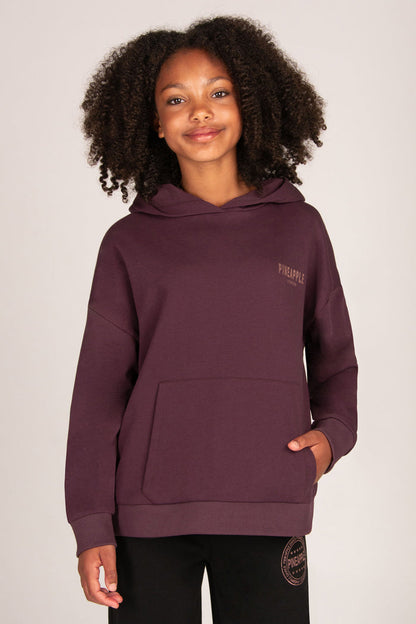A smiling young girl wearing a dark plum purple pullover hoodie with a small rose-gold 'PINEAPPLE' logo on the left chest, paired with black trousers, against a solid light grey background.