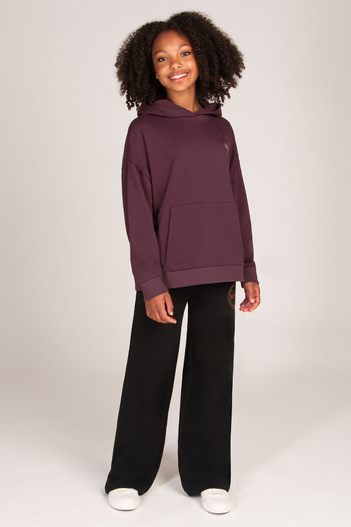 A young girl stands against a light grey studio background wearing a dark aubergine pullover hoodie, black wide-leg sweatpants, and white sneakers.