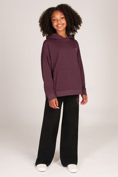 A young girl stands against a light grey studio background wearing a dark aubergine pullover hoodie, black wide-leg sweatpants, and white sneakers.