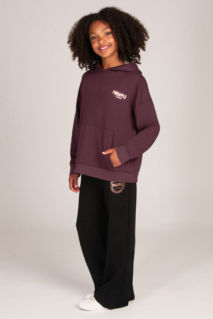A young girl stands against a light grey studio background wearing a dark aubergine pullover hoodie, black wide-leg sweatpants, and white sneakers.
