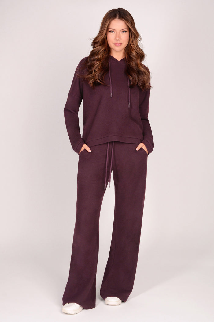 Woman wearing maroon brown wide leg trousers with matching dip hem hoodie on a plain background.