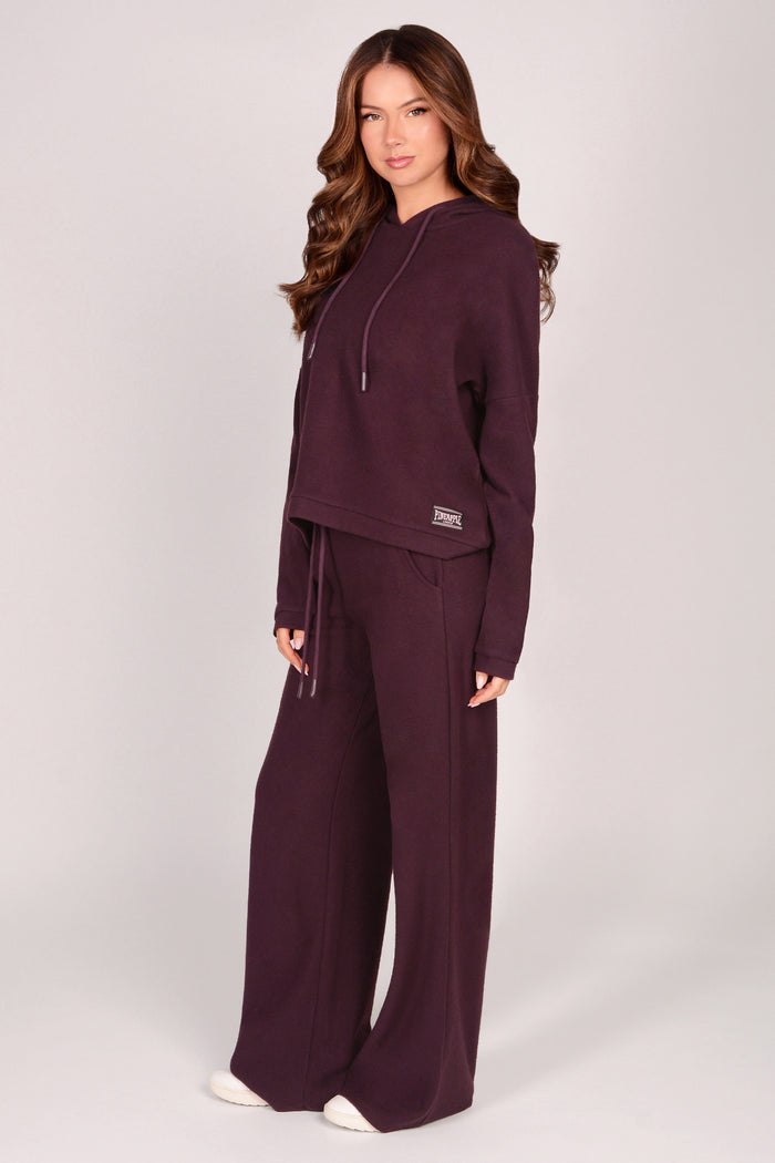 Woman wearing maroon brown wide leg trousers with matching dip hem hoodie on a plain background.