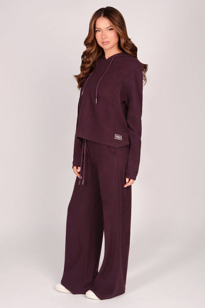Woman wearing maroon brown wide leg trousers with matching dip hem hoodie on a plain background.