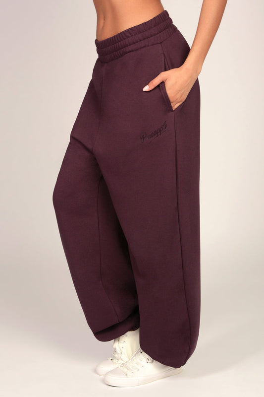 A person wearing loose, wide-leg jogger sweatpants in a deep plum purple color, paired with white sneakers, shown from the waist down against a light grey studio background.