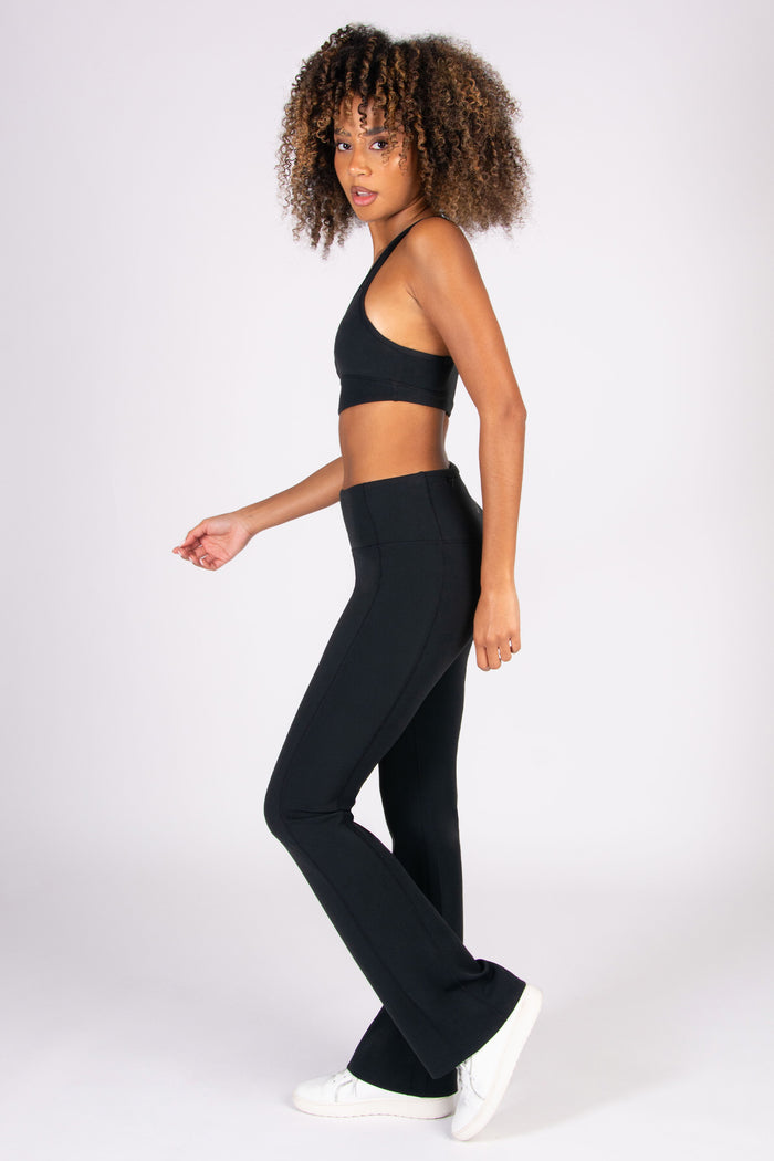 Woman wearing a black sports bra and matching black super soft yoga flared trousers on a white background