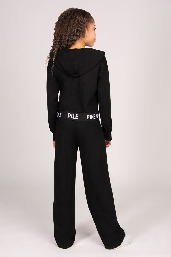 Girl wearing a black hoodie with matching trousers against a plain grey background.