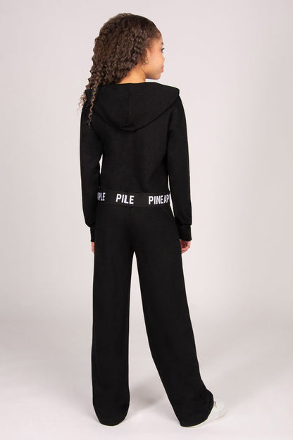Girl wearing a black hoodie with matching trousers against a plain grey background.