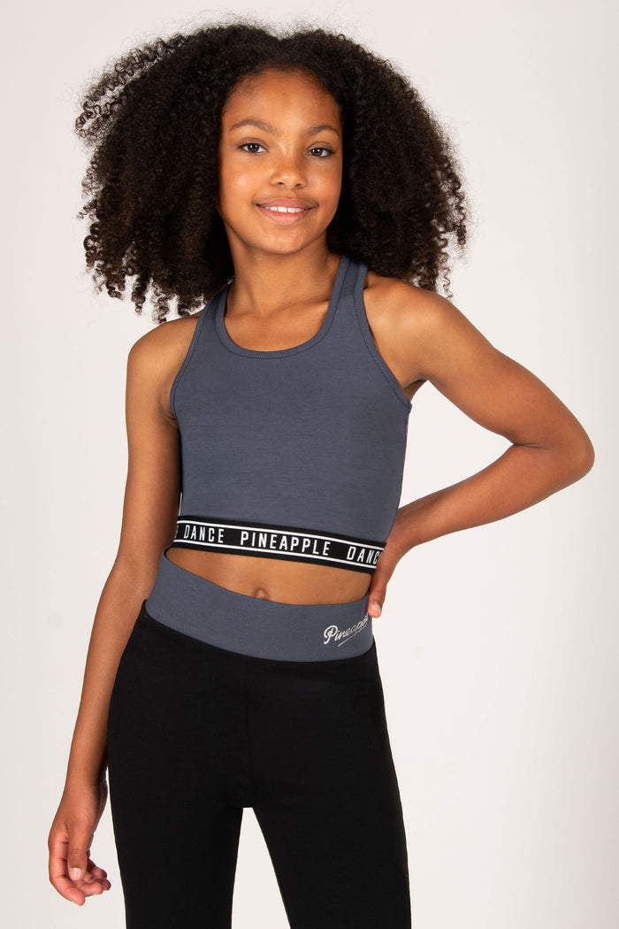 Young girl wearing a dark grey sports bra and black leggings with 'Pineapple' branding on a white background