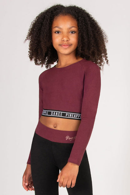 Young girl wearing a maroon long-sleeve crop top with black tape waistband on a plain background