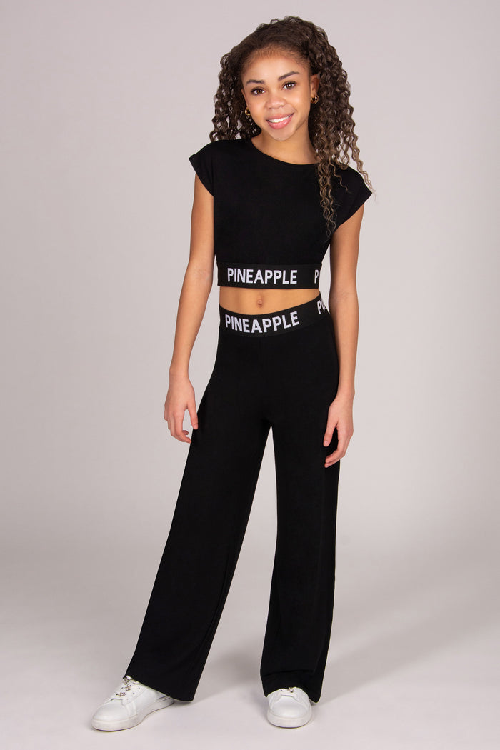 Young girl wearing a black 'Pineapple' branded crop top and matching wide leg trouserson a plain background